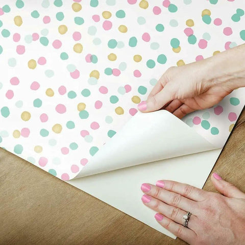 Hands peeling Pink And Blue Glitter Confetti Peel And Stick Wallpaper Sample, showing repositionable and removable feature on wood surface.