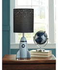 Kids Rocket Table Lamp With Gray Ceramic Base And Fabric Drum Shade on wooden desk beside globe and books by window.