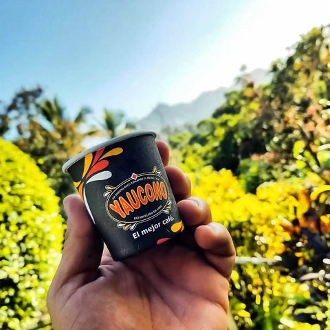 A hand holding a Yaucono espresso cup in a tropical outdoor setting with lush greenery and mountains.