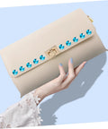 Front view of beige clutch bag with a row of turquoise metal studs along the flap and a gold clasp.