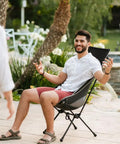 Man sitting on Chillin Chair Ultra Lightweight Foldable Portable Outdoor Seat For Tailgating Camping Backyard Barbecues And More Comfortable Durable Easy Setup Go To Seat For Family Trips Picnics And Adventures by a poolside.