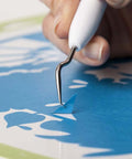 Cricut Basic Tool Set: close-up of white plastic-handled precision weeding tool with curved metal tip on blue vinyl.