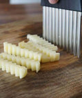 Kakalote crinkle cutter with black handle and stainless steel blades slicing potatoes into wavy fries on a wooden board.