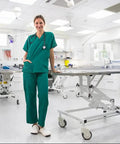 Medical professional in scrubs standing in a clean, modern hospital examination room with medical equipment and adjustable table.
