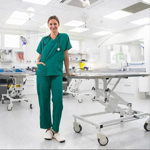 Medical professional in scrubs standing in a clean, modern hospital examination room with medical equipment and adjustable table.