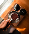 Hand holding portafilter with grounds next to 58mm Coffee Distribution Tool For Espresso Portafilter Leveler With Adjustable Depth And Stainless Steel Base Barista Leveling Stand For Even Grounds And Consistent Tamp Go To Accessory For Home Coffee Enthusiasts