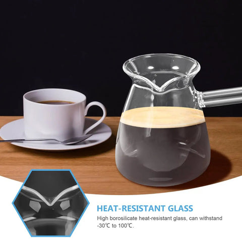 Glass coffee pot with handle on wooden table, alongside white cup and saucer. Close-up of heat-resistant glass feature.