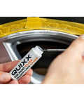 Hand holding QUUIXX rim paint bottle preparing to touch up black rim as part of Solid Alloy Wheel Repair Kit For Black Rims Smooths Out Curb Rash And Road Scuffs DIY Two Part Liquid Aluminum Compound Plus Spot On Black Touch Up Paint For A Fresh New Ride Glow Up.
