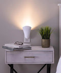 Plug In Wall Sconce Uplighter With GU10 Spotlight Wash Light on bedroom wall, casting warm glow on nightstand with books and plant, no LED bulb included.
