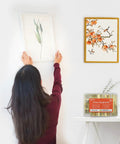 Woman hanging a framed plant artwork on a wall using the Rock Solid Picture Hanging Kit Top Shelf Heavy Duty Hooks And Nails For Drywall Wood No Sweat Setup For Photo Frames Mirrors And Wall Art 152 Piece Set.