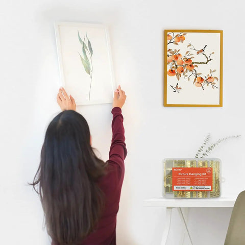 Woman hanging a framed plant artwork on a wall using the Rock Solid Picture Hanging Kit Top Shelf Heavy Duty Hooks And Nails For Drywall Wood No Sweat Setup For Photo Frames Mirrors And Wall Art 152 Piece Set.