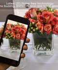 Smartphone capturing a photo of a vibrant floral arrangement in a clear glass vase with LED lighting in background.