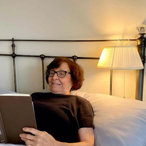 Over The Bed Reading Lamp With Soft Cream Pleated Fabric Shade And No Fuss Easy On Off Pull Cord Eye Friendly Bedside Light Great For Reading Knitting Puzzles And Late Night Chats In Bed Mounts Above The Headboard illuminating a woman reading in bed