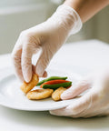 Hand wearing Top Shelf Disposable Clear Vinyl Exam Gloves Medical Grade XL 100 Count Pack grabbing food on a plate.
