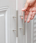Hand approaching stainless steel T bar cabinet handles with satin brushed nickel finish, installed on white cupboard doors.