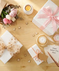 A Mother's Day gift setup with flowers, candles, and cards on a wooden table, featuring white boxes with pink bows.