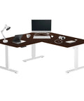 L shaped electric standing desk with 60x60 inch walnut surface, white frame, laptop, and lamp; perfect for home office.