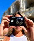 A Real Deal 16 Megapixel Digital Camera With 5X Optical Zoom being held up for a selfie in front of a historic building.