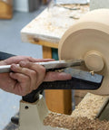 Carbide woodturning tool on a lathe shaping a spinning wooden blank; hands guide the tool along the rest.