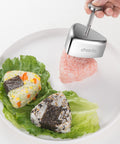 Choxila stainless steel onigiri mold with spring press; two triangular rice balls on lettuce, one wrapped in nori.