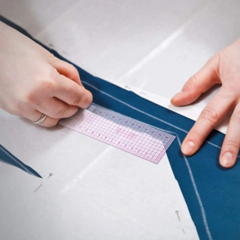 Hands using a pink grid ruler on blue fabric while drafting a sewing pattern with the Top Shelf 13 Piece French Curve Ruler Set For Sewing Pattern Drafting And Fashion Design Includes Hip Curves Tracing Wheels And Measuring Tape Ace Gear To Nail Your Next Creative Project.