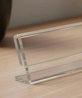 Close-up of a clear acrylic desk name plate with a horizontal slant back, part of a 12 pack 2x8 sign holders, on a wooden table.