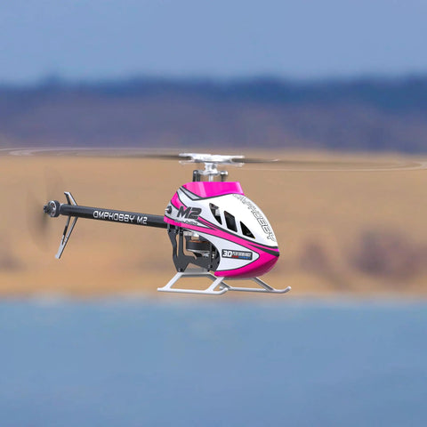 M2 V3 Sport BNF RC Helicopter in flight, pink body with OFS3 Plus Flight Controller, over a scenic outdoor backdrop