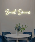 Lit Neon Sign Wall Art Decor 25x6.4 Inches Real LED Neon Light For Bedroom Office Dorm Farmhouse Vibe Aesthetic Room Decor Valentine Party Hanging Sign "Sweet Dreams" mounted above a round table with flowers.