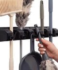 Heavy Duty Broom And Mop Wall Mount Organizer With Five Slots And Six Hooks Wall Mounted Broom Rack For Brooms Mops And Cleaning Tools No Fuss Space Saving Storage Solution For Kitchen Garage Laundry And Garden, holding a broom and cleaning tools.