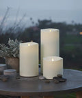 Outdoor Moving Flame Pillar Candle With Remote Included, IPX4 weatherproof LED, ivory 6x6, on patio table with plants.