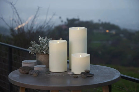 Outdoor Moving Flame Pillar Candle With Remote Included, IPX4 weatherproof LED, ivory 6x6, on patio table with plants.
