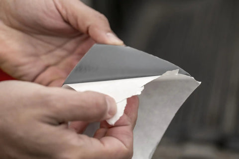 Hands holding Heavy Duty Waterproof Repair Tape, peeling backing, showing adhesive side. 2 inch by 5 foot roll in black color.