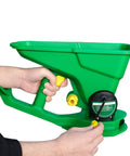 Hands holding a green cordless handheld seed and fertilizer spreader with adjustable feed rate, portable for lawns and gardens.