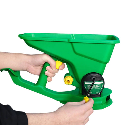 Hands holding a green cordless handheld seed and fertilizer spreader with adjustable feed rate, portable for lawns and gardens.