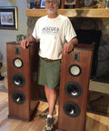 Man standing next to two wooden tower speakers in a living room setting, showcasing the speaker design and size.