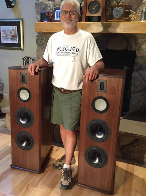 Man standing next to two wooden tower speakers in a living room setting, showcasing the speaker design and size.
