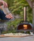 Person seasoning pizza dough near gas burner attachment for Karu 2 Pro pizza oven - 28mbar propane, includes regulator and hose, converter kit.