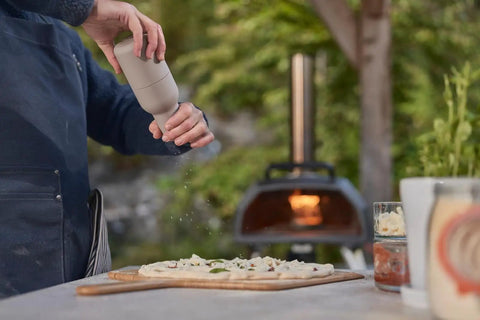 Person seasoning pizza dough near gas burner attachment for Karu 2 Pro pizza oven - 28mbar propane, includes regulator and hose, converter kit.