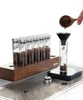 12 Piece Single Dose Coffee Bean Storage Tubes With One Way Exhaust Valve Glass Vials On Sturdy Walnut Wood Base pouring beans.
