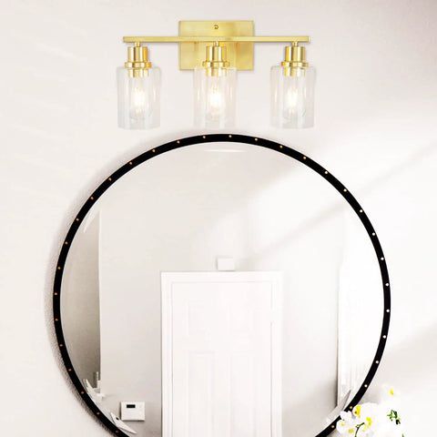 Three Light Sleek Gold Brushed Brass Vanity Light Fixture With Clear Glass Shade For Modern Bath Wall Hallway Bedroom And Living Room Bulbs Not Included shown above a round mirror on a wall.