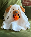 Stuffed animal with a white ghost costume holding a pumpkin bucket on a couch.