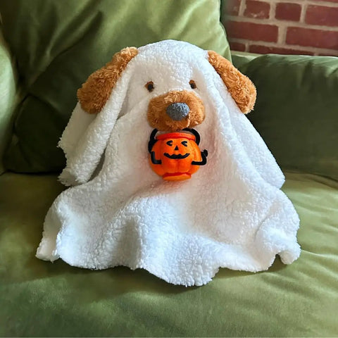 Stuffed animal with a white ghost costume holding a pumpkin bucket on a couch.
