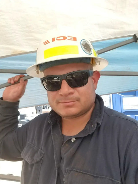 Man wearing Men's Safety Glasses With Smoke Lens, Matte Black Wraparound Frame, ANSI Z87 compliant sunglasses under a work hat.