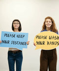 Two women holding signs reading 'Please Keep Your Distance' and 'Please Wash Your Hands' with a neutral background.
