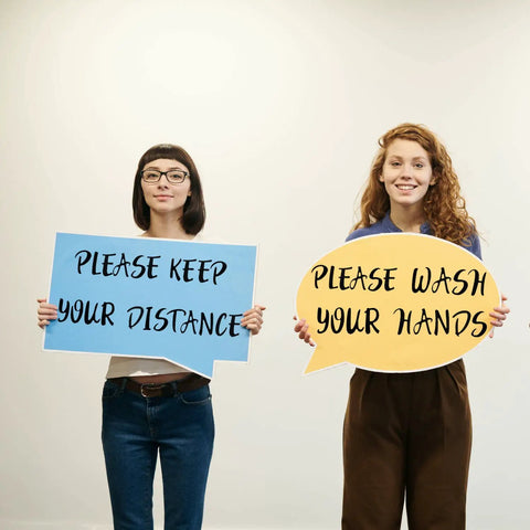 Two women holding signs reading 'Please Keep Your Distance' and 'Please Wash Your Hands' with a neutral background.