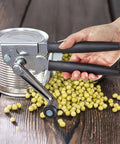 Heavy Duty Manual Can Opener With Rubber Coated Long Handles, Commercial Grade Squeeze And Crank Can Opener For Kitchen, Restaurant, And Outdoor Picnics, Durable Stainless Steel Body, Easy Grip, Long Lasting Performance in use opening a can on wooden table.
