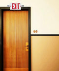 Red LED Exit Sign With Emergency Lights And Battery Backup, Dual Adjustable Head LED Lamps, above a wooden door in hallway.