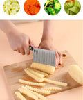 Side view of a stainless steel crinkle cutter knife with a black handle slicing potato sticks on a wooden cutting board