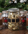 Five mason jars decorated as reindeer with googly eyes and red nose, filled with candies, set on a wooden table with holiday decor.