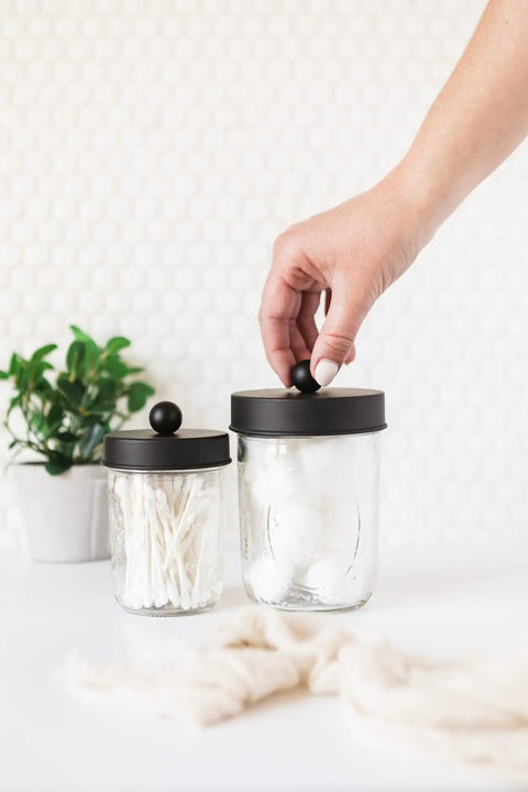 Hand opening Apothecary Lid Storage Set With Ball Mason Jars Two Pack Farmhouse Style Vanity Organizer For Kitchen Bath Office Black Finish Totally Game Changing Storage Jars For Countertop Organization, with plant in background